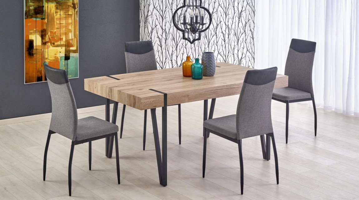 Stół na metalowych nogach u boku krzeseł tapicerowanych tkaniną z modnym obrazem na szarej ścianie