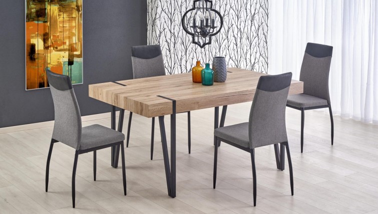 Stół na metalowych nogach u boku krzeseł tapicerowanych tkaniną z modnym obrazem na szarej ścianie