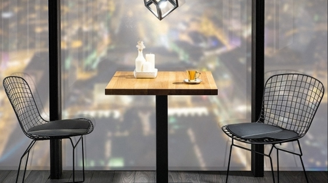 Drewniany stół na jednej nodze z kwadratowym blatem w zestawieniu z metalowymi krzesłami w jadani na tle dużego okna